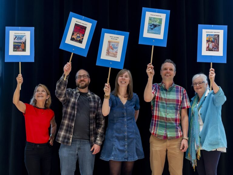 Authors Wanda Baxter, Braden Hallett, Heidi Tattrie Rushton, Pheilm Martin, and Margaret MacKay stand together holding blue signs displaying their book covers and names and smiling broadly.