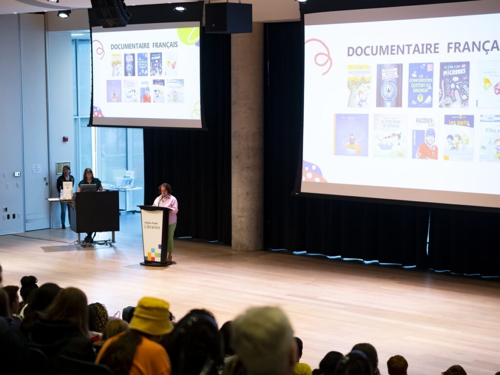 Big screens display French nonfiction nominees above a podium with a white woman wearing a pink shirt standing at the microphone. There's a crowd of kids and chaperones in the foreground of the photo.