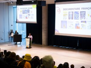 Big screens display French nonfiction nominees above a podium with a white woman wearing a pink shirt standing at the microphone. There's a crowd of kids and chaperones in the foreground of the photo.