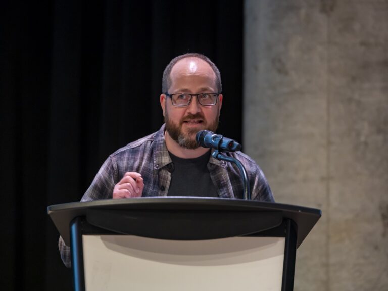 White man with short dark hair, beard, and dark-rimmed glasses wearing a grey plaid shirt over a black t-shirt speaks at a podium.