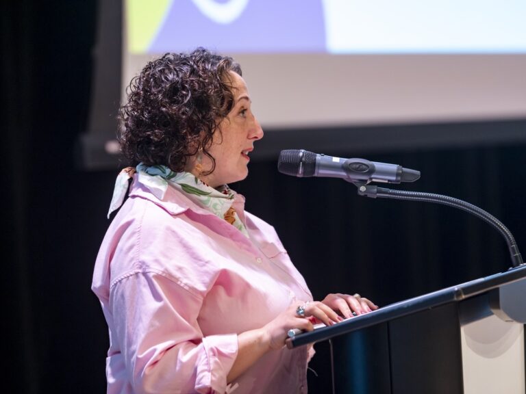 A white woman wearing a pink shirt stands at a podium with a microphone.