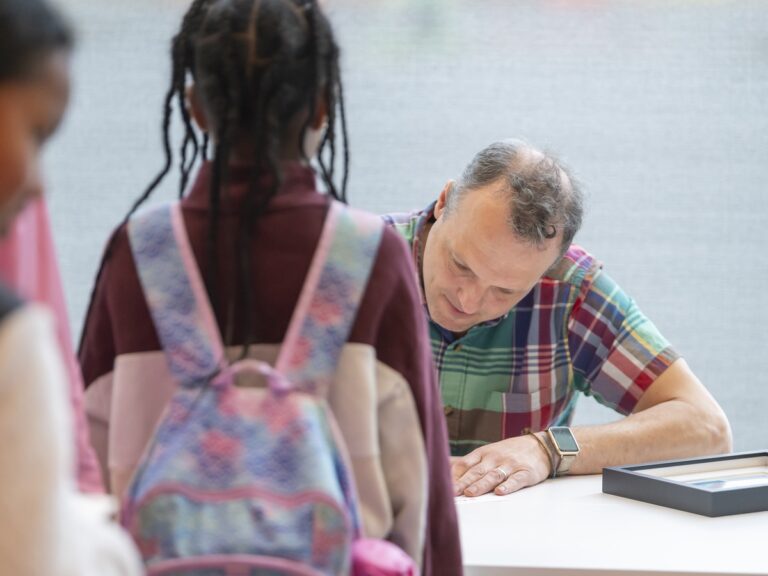 Pheilm Martin, a white man with short curly salt-and-pepper hair wearing a green, red, blue, and white plaid shirt sits at a white table signing autographs for a student whose back is to the camera. Next to Pheilm's elbow is a framed Hackmatack certificate.
