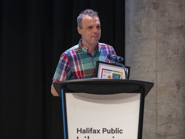 White man with short curly salt-and-pepper hair wearing a green, red, blue, and white plaid shirt holds a framed certificate as he stands at a podium.