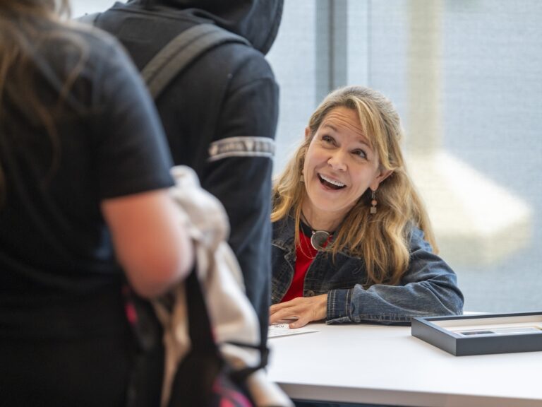 Wanda Baxter, a white woman with long light brown hair wearing a denim jacket over a red top, smiles as she signs autographs for students. There are two students visible in the lineup wit their backs to the camera. A framed certificate sits on the white table next to Wanda.