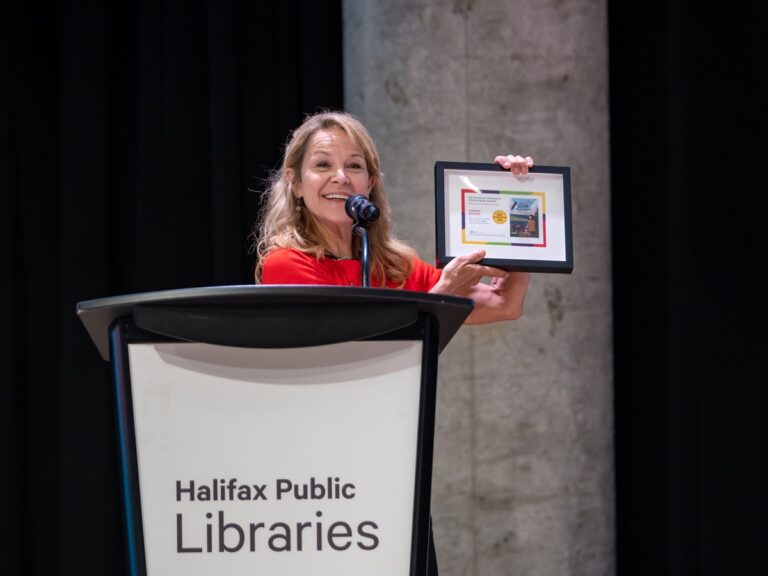 Wanda Baxter, a white woman with long light brown hair and red top, holds a framed certificate as she smiles while standing at a podium.
